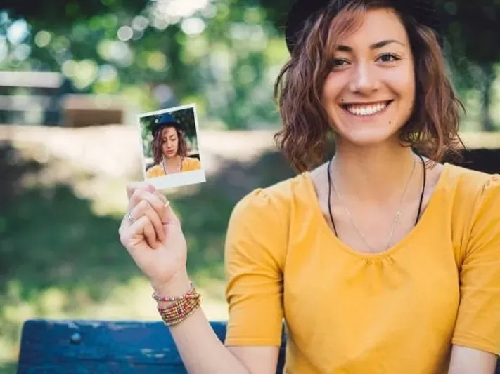 A smiling woman in a yellow top and a black hat, sitting on a park bench